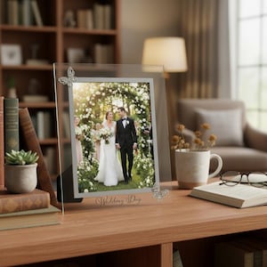 Puede incluir: Un marco de fotos de vidrio transparente con una foto de boda. El marco tiene adornos de mariposas plateadas y la inscripción "Wedding Day". El marco se exhibe sobre una superficie de madera.