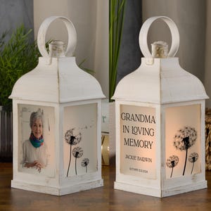 Op de afbeelding: Witte lantaarn met een foto van een vrouw en een paardenbloemdessin. De lantaarn heeft de tekst "Grandma in loving memory Jackie Baldwin 11.07.1955-12.02.2024".