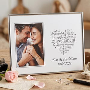 Könnte beinhalten: Silberner Verlobungs-Fotorahmen mit weißem Passepartout. Der Rahmen fasst ein Foto von 10 cm x 15 cm und enthält den Text "We're Engaged" und "Jan & La Verne 18.05.2026".