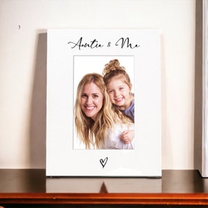 Puede incluir: Marco de fotos blanco con una inscripción negra "Auntie & Me" y un símbolo de corazón. El marco contiene una foto de una mujer adulta y una niña sonriendo.