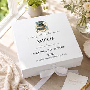 Può includere: Una scatola regalo quadrata bianca con chiusura a nastro, caratterizzata da un'illustrazione ad acquerello di un tocco di laurea su dei libri. Il testo recita: congratulations AMELIA on Your Graduation UNIVERSITY OF LONDON 2026 BA TOWN AND COUNTRY PLANNING.