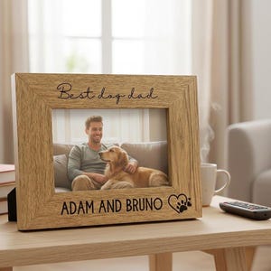 A wooden picture frame with a photo of a man and a dog, along with the text "Best dog dad" and "Adam and Bruno" engraved on it, sits on a wooden table in a living room.