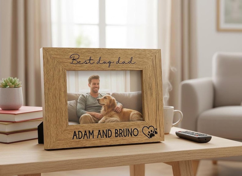 A wooden picture frame with a photo of a man and a dog, along with the text "Best dog dad" and "Adam and Bruno" engraved on it, sits on a wooden table in a living room.