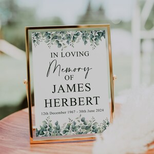 May include: A framed memorial sign with a white background and green foliage border. The text reads "In Loving Memory of James Herbert 12th December 1967 - 30th June 2024".