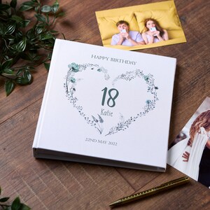 May include: White hardback photo album with a floral heart design. The heart has the number 18 and the name Katie inside. The text "Happy Birthday" and "22nd May 2022" are printed on the cover.