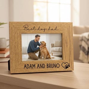 A wooden picture frame with a photo of a man and a dog inside, placed on a wooden surface. The frame has a personalized message that reads "Best day dad" and "Adam and Bruno".