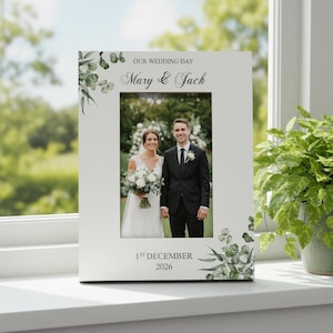 Può includere: Una cornice bianca con la scritta "OUR WEDDING DAY" e i nomi "Mary & Jack". Contiene una foto di matrimonio di una coppia. La data "1st December 2026" è stampata in basso. Accenti di verde decorano gli angoli.
