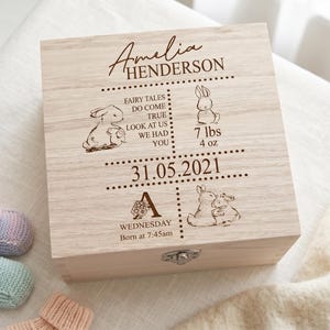 Può includere: Una scatola ricordo in legno chiaro con il nome "Amelia Henderson" inciso sul coperchio. La scatola presenta illustrazioni di conigli, la data 31.05.2021 e il testo "FAIRY TALES DO COME TRUE LOOK AT US WE HAD YOU". Include anche il peso del bambino: 3,18 kg.