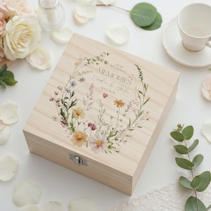 May include: A light-coloured wooden box with a hinged lid. The lid features a floral wreath design and the words "OUR WEDDING MEMORIES", a couple's names and a date. The box has a metal clasp.