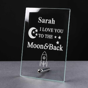 May include: A glass plaque with a white etched design of a crescent moon, stars, and a rocket ship. The text reads "Sarah I love you to the moon & back".