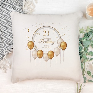 May include: White throw pillow with a gold circle design featuring the number 21, the words "Happy Birthday", a name, and the date 14-06-2022. The circle is surrounded by gold confetti and gold balloons with white strings.