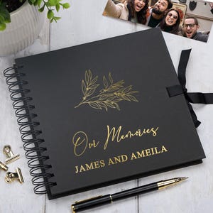 May include: A black photo album with a spiral binding, gold lettering, and a botanical illustration. The cover says "Our Memories" and includes names. A black ribbon and pen are also present.