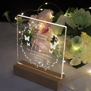 K&ouml;nnte beinhalten: Ein wei&szlig;es, beleuchtetes Acrylschild mit einem Blumenkranz und Schmetterlingen. Das Schild tr&auml;gt die Aufschrift "In liebevoller Erinnerung an eine wundervolle Mama. Du bist das Licht, das f&uuml;r immer in unseren Herzen leuchtet. Wir vermissen dich 1944-2021".