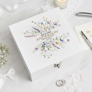 May include: A white wooden memory box featuring a floral design and the text "MR & MRS ANDERSON WEDDING MEMORIES 21.06.2036". The box has a silver clasp and two silver rings are visible.
