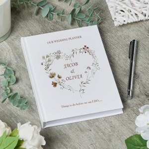 Puede incluir: Planificador de bodas blanco con un dise&ntilde;o de coraz&oacute;n floral y los nombres Jacob & Olivia. El texto "Our Wedding Planner" y "Things to do before we say I DO..." est&aacute; impreso en la portada.
