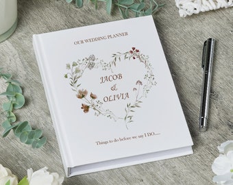Può includere: Un planner di nozze bianco con un design a forma di cuore floreale e i nomi Jacob & Olivia. Il testo "Our Wedding Planner" e "Things to do before we say I DO..." è stampato sulla copertina.