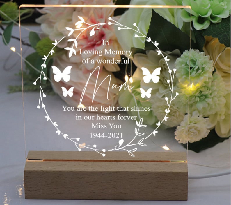 K&ouml;nnte beinhalten: Ein wei&szlig;es, beleuchtetes Acrylschild mit einem Blumengirlanden und Schmetterlingen. Das Schild tr&auml;gt die Aufschrift "In Loving Memory of a wonderful Mum You are the light that shines in our hearts forever Miss You 1944-2021".