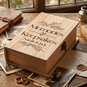 May include: A rectangular light-coloured wooden box with a hinged lid featuring a dark engraved design. The text reads Memories & Keepsakes with the names Natalie & Jack, accompanied by leaf illustrations. A metal latch secures the side of the box.