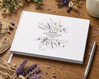 May include: A white rectangular wedding guest book with a floral illustration of wildflowers in shades of blue, purple, and yellow. The text reads MR & MRS ANDERSON WEDDING GUEST BOOK 23.06.2026. The book is positioned on a wooden surface with a silver pen.