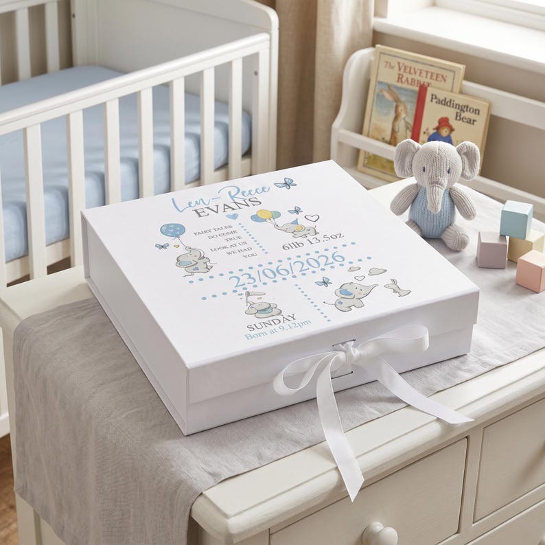 A white baby box with a ribbon sits on a wooden dresser, next to a crib and a shelf with books.