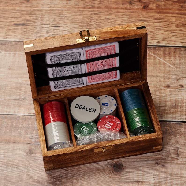 May include: A wooden box with a hinged lid, containing two decks of playing cards and a variety of poker chips. The box is open, revealing the contents. The chips are in various colors, including red, green, and blue. The box is labeled "DEALER" on one of the chips.