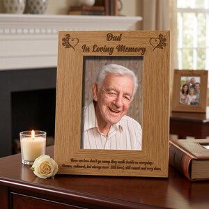 a wooden picture frame with a photo of an elderly man, placed on a wooden desk. The frame has a message engraved on it that reads "Dad in Loving Memory". Next to the frame, there is a lit candle and a book.