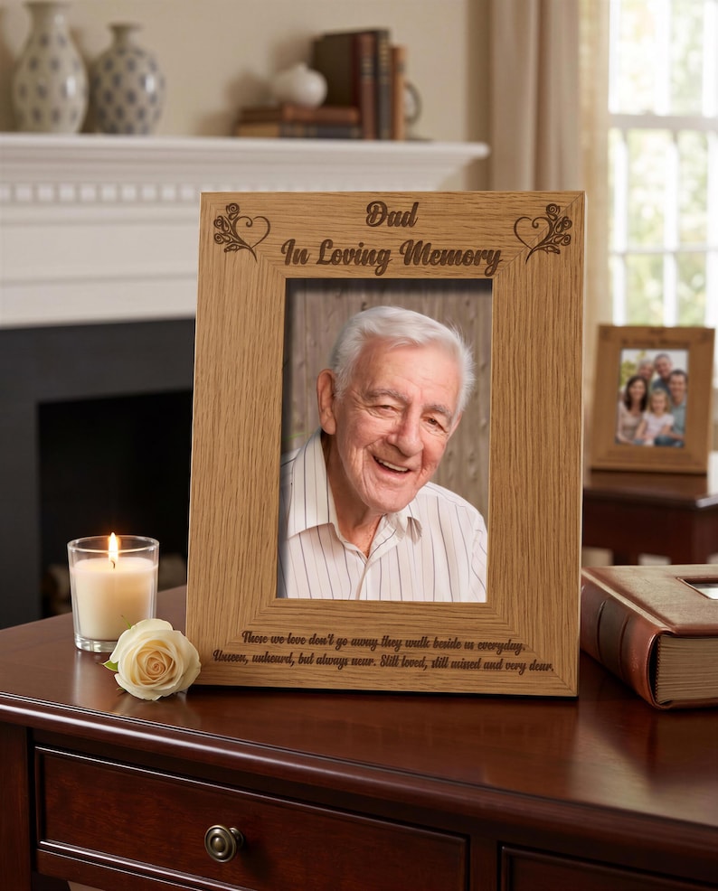 a wooden picture frame with a photo of an elderly man, placed on a wooden desk. The frame has a message engraved on it that reads "Dad in Loving Memory". Next to the frame, there is a lit candle and a book.