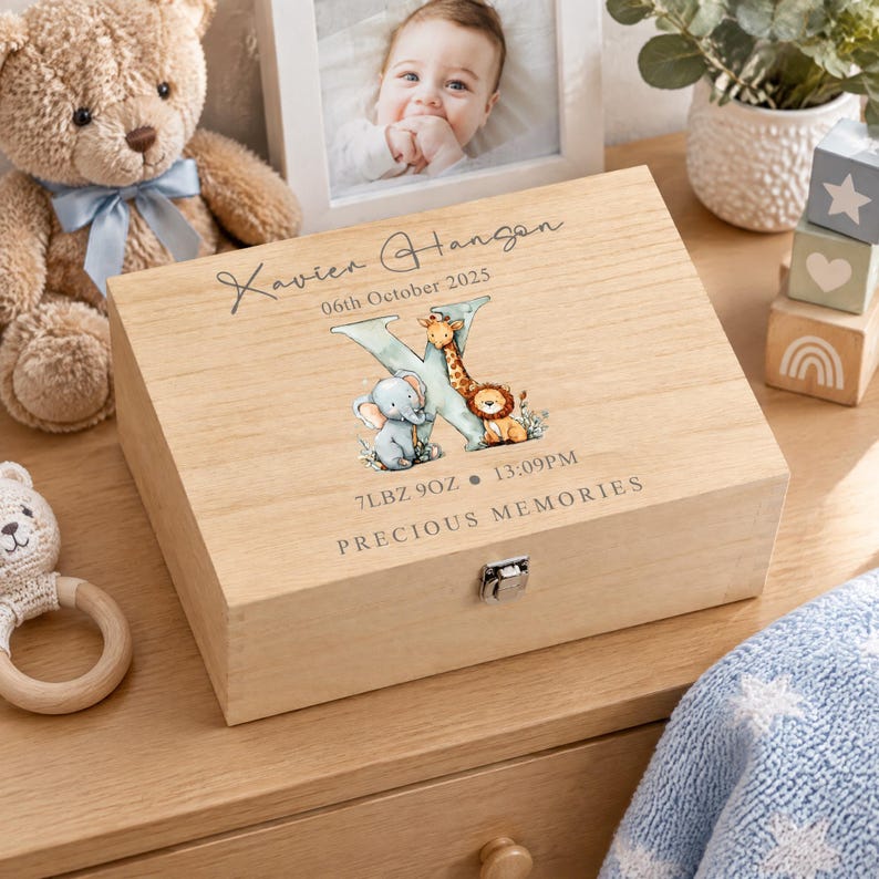 A wooden box with a personalized name and date, featuring a baby's photo and an illustration of animals, sits on a wooden surface surrounded by various baby-related items.