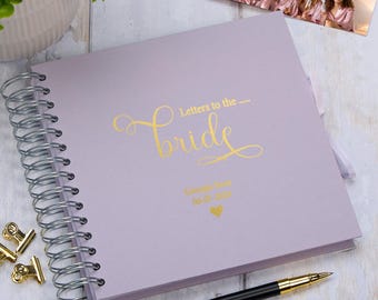 Op de afbeelding: Een lavendelkleurig spiraalgebonden notitieboek met gouden folielettering die "Brieven aan de... Bruid" en een gouden hartje zegt. Het notitieboek is gepersonaliseerd met de tekst "Gastenboek" en een datum.