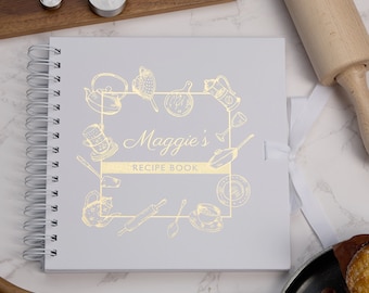 May include: A white recipe book with a gold foil design of kitchen utensils and the text "Maggie's Recipe Book".
