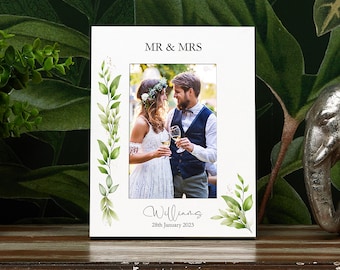 May include: A white picture frame with the words "MR & MRS" at the top and the name "Williams" and the date "28th January 2023" at the bottom. The frame has a photo of a couple and is decorated with green leaves.