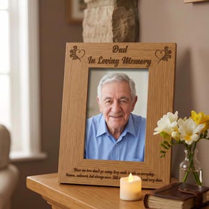 a wooden picture frame with a photograph of an older man, placed on a wooden table. The frame has a message engraved on it that reads "Dad in Loving Memory". Next to the frame, there is a vase with white flowers, and a lit candle.