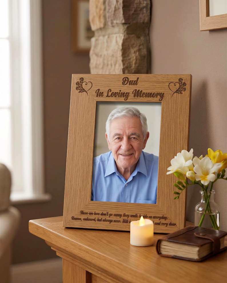 a wooden picture frame with a photograph of an older man, placed on a wooden table. The frame has a message engraved on it that reads "Dad in Loving Memory". Next to the frame, there is a vase with white flowers, and a lit candle.