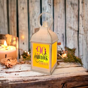 Op de afbeelding: Een witte lantaarn met een geel en roze ontwerp met de tekst "90 & Fabulous" en daaronder de naam Amelia. De lantaarn is verlicht en heeft een warme gloed.