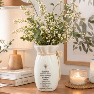 May include: A white ceramic vase with a cinched neck tied with a beige rope. The vase features a small floral illustration and text that reads: Amanda, No matter where life takes us, or how far apart we may be, you'll always mean so much to me.