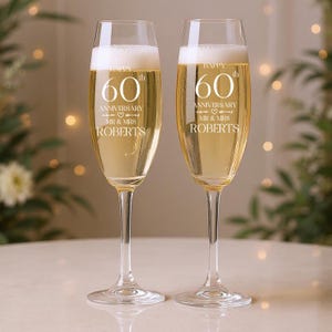 May include: Two champagne flutes filled with a golden liquid. The glasses are engraved with "Happy 60th Anniversary" and the name "Roberts". The flutes are on a white surface, with a blurred background of greenery and lights.