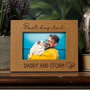 A wooden picture frame with a photo of a man in a yellow sweater hugging a small white dog. The frame has the text "Best dog dad" and "DADY AND STORM" engraved on it.