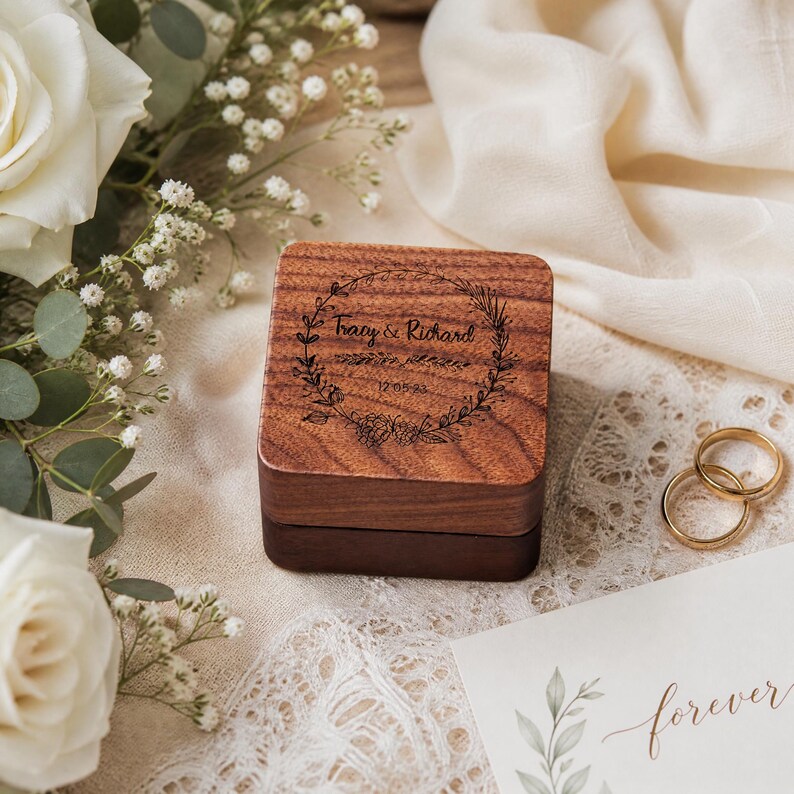 A wooden box with a floral design, surrounded by white roses, greenery, and a lace doily, with a wedding invitation card and a pair of gold wedding rings.