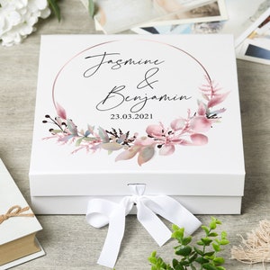 May include: White gift box with a pink and green floral wreath and a gold circle around the text "Jasmine & Benjamin 23.03.2021".