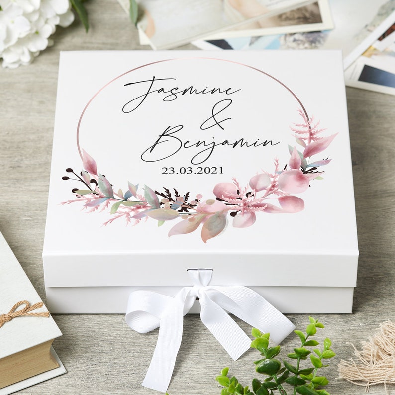 May include: White gift box with a pink and green floral wreath and a gold circle around the text "Jasmine & Benjamin 23.03.2021".