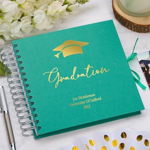 May include: A teal green graduation guest book with a gold foil graduation cap and the word "Graduation" in gold foil lettering. The book has a gold ribbon and a spiral binding. The book is personalised with the name "Joe Henderson", "University of Salford", and "2022".