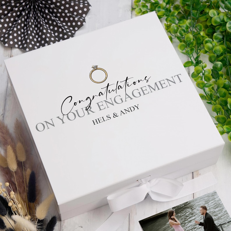 A white box with a gold ring on it, placed on a wooden surface. The box has text that reads "Congratulations ON YOUR ENGAGEMENT HELS & ANDY".