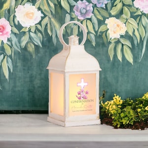 May include: A white metal lantern with a handle, featuring a glowing panel with a cross and floral design. The text "Confirmation of Amanda Smith" is visible. The lantern is set against a backdrop of painted flowers and foliage.