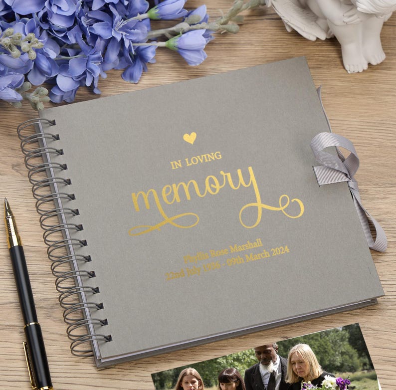 May include: Gray spiral bound memory book with a gold foil heart and the words "In Loving Memory" in a gold script font. The book has a gray ribbon closure and the text "Phyllis Rose Marshall Died May 20th, 2024" is printed in gold foil.