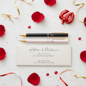 May include: Two pens, one black and one pale pink, both with gold detailing and the number "40", lie above a white case with the inscription "Adam & Melanie" and "40 years together". Red petals and a "40" balloon are scattered around.