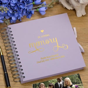 May include: A lavender-colored memory book with a gold heart and the words "In Loving Memory" in gold script. The book has a spiral binding and a gold ribbon. The book is personalized with the name "Phyllis Rose Marshall" and the dates "22nd July 1936 - 09th March 2021".