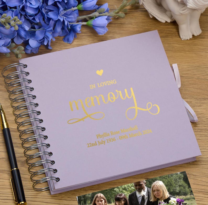 May include: A lavender-colored memory book with a gold heart and the words "In Loving Memory" in gold script. The book has a spiral binding and a gold ribbon. The book is personalized with the name "Phyllis Rose Marshall" and the dates "22nd July 1936 - 09th March 2021".