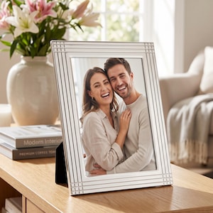May include: A rectangular silver-toned picture frame with a vertical and horizontal grooved border pattern. The frame holds a photograph of a smiling couple embracing. It features a black easel-style back support for standing on a flat surface.