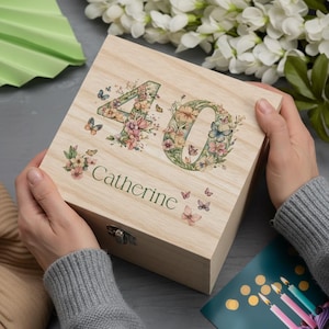 May include: A wooden box featuring a floral "40" design and the name "Catherine". The numbers and butterflies are decorated with pink, yellow, and blue flowers. The box has a metal clasp and is held by a person wearing a grey jumper.