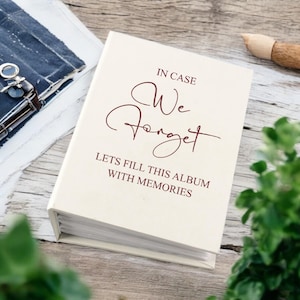 Puede incluir: Un álbum de fotos blanco con el texto "En caso de que olvidemos, llenemos este álbum con recuerdos" impreso en la portada.